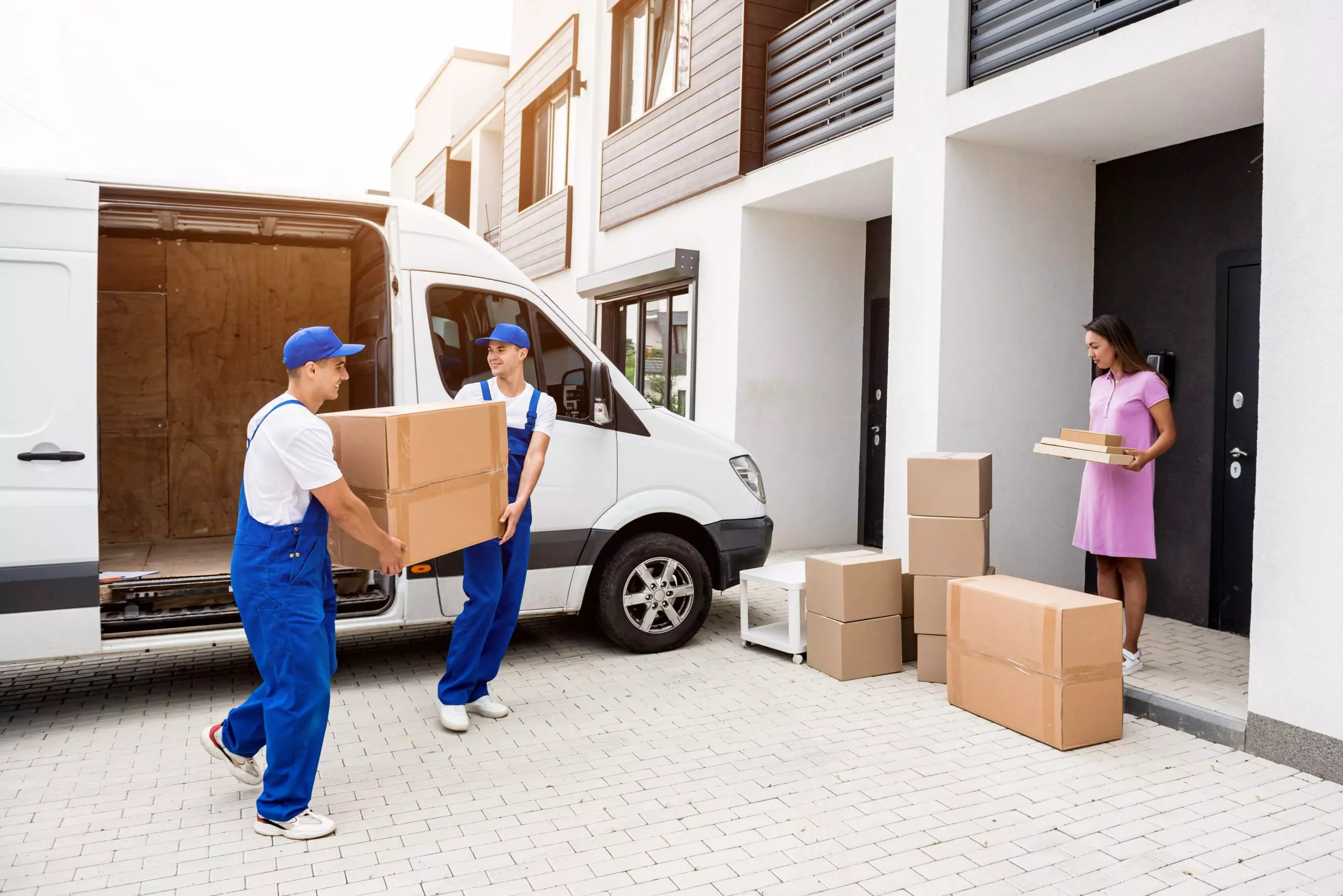 Zwei Umzugshelfer in blauen Uniformen eines Umzugsunternehmens laden Kisten aus einem weißen Lieferwagen aus. Umzugsfirma-SUMO Entrümpelung