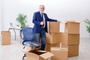 Ein Mann im Anzug streckt den Daumen nach oben, während er neben mehreren Kartons eines bw Umzugsunternehmens in einem spärlich möblierten Büroraum steht.
