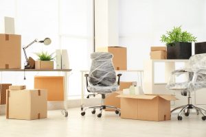 Ein Büroraum mit zwei in Plastik eingewickelten Stühlen, Umzugskartons der Umzugsfirma BW, einer Schreibtischlampe und Topfpflanzen.