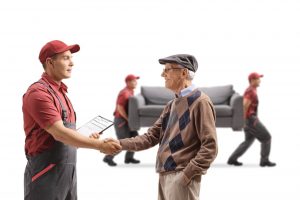 Ein Lieferant in Uniform schüttelt einem älteren Herrn die Hand, im Hintergrund transportieren zwei Möbelpacker der Umzugsfirma ein Sofa.