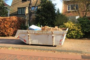 Ein großer weißer Müllcontainer voller Kartons und anderem Abfall steht am Straßenrand in einem von Bäumen und Häusern umgebenen Wohngebiet.