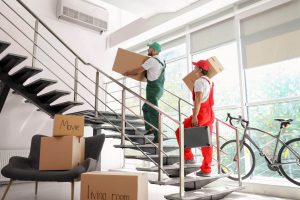 Zwei Arbeiter in Overalls der Umzugsfirma tragen Kisten und Ausrüstung eine moderne Treppe hinauf. Kisten mit der Aufschrift „Kino“ und „Wohnzimmer“ stehen neben einem Fahrrad und einem Stuhl in einem lichtdurchfluteten Raum.
