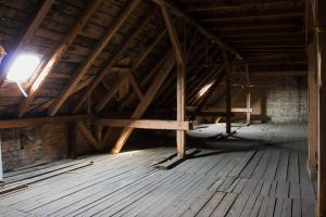 Ein leerer, schwach beleuchteter Dachboden mit freiliegenden Holzbalken, Holzboden und Licht, das durch ein kleines Fenster hereinfällt.